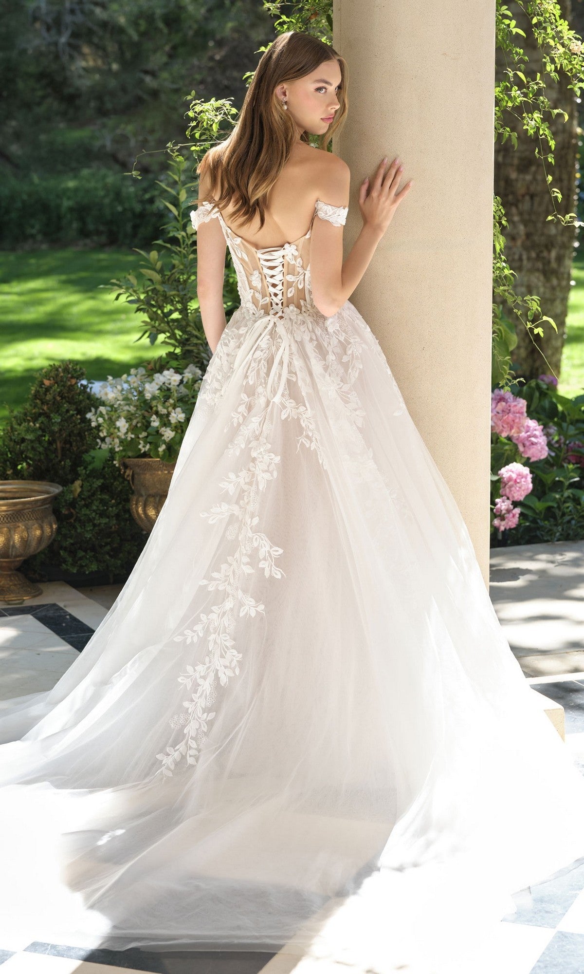 A woman wears the Andrea and Leo WL057 long wedding dress with a lace-up back and floral details, standing outdoors near a column and garden, facing away.